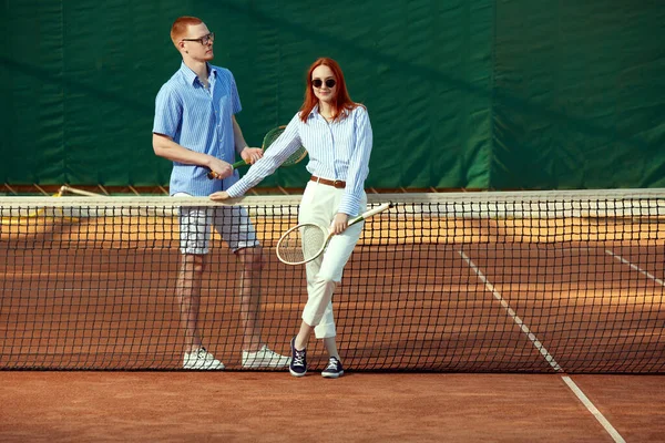 年轻美丽的红头发女子和英俊的男子穿着时髦的休闲装和时髦的眼镜配件在网球场上摆设 体育概念 积极的生活方式 业余爱好 闲暇时间 — 图库照片