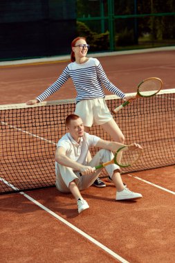 Gündüz vakti açık tenis kortunda poz veren genç ve güzel bir kadın ve şık giyinen yakışıklı bir adam. Gelişigüzel moda, spor, aktif yaşam tarzı, boş zaman, reklam