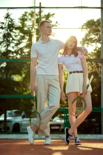 美丽的年轻夫妇 红头发的男人和女人穿着时髦的白色衣服 日落时在球场上摆着网球拍 休闲时尚 积极的生活方式 业余爱好 闲暇时间 广告的概念 — 图库照片