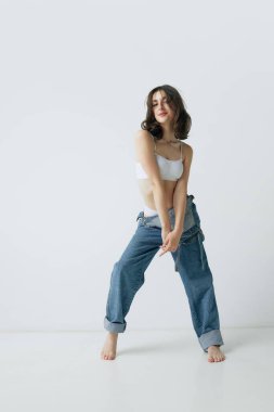 Güzel, kıvırcık, ince vücutlu, kot pantolonlu ve gri stüdyo arka planında poz veren genç bir kadının tam boy portresi. Güzellik kavramı, vücut ve cilt bakımı, sağlık, sağlık, sağlık, reklâm