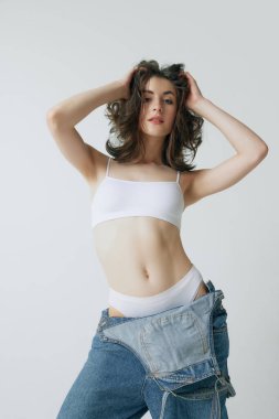 Kıvırcık saçlı, zayıf vücutlu, gri stüdyo arka planında kot ve üstte poz veren güzel esmer kadın. Güzellik kavramı, vücut ve cilt bakımı, sağlık, sağlık, sağlık, reklâm