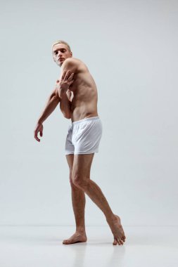 Sarı saçlı, kaslı vücutlu, gri stüdyo arka planında iç çamaşırlarıyla poz veren yakışıklı bir gencin tam boy portresi. Erkek doğal güzellik kavramı, vücut bakımı, sağlık, spor, reklam