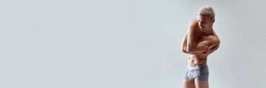 Yakışıklı, sağlıklı, güçlü, kaslı vücutlu, gri stüdyo arka planında iç çamaşırsız poz veren genç bir adam. Erkek doğal güzellik, vücut bakımı, sağlık, spor, moda, reklam, heykel sanatı kavramı