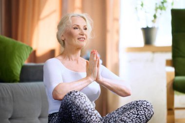 Güzel, güler yüzlü, şık spor kıyafetleri içinde sabahları evde yoga egzersizleri yapan son sınıf kadını. Sportif ve sağlıklı yaşam tarzı kavramı, yaş, vücut bakımı, spor, sağlık.