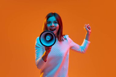 Günlük kıyafetli genç bir kadın megafonla turuncu stüdyonun arka planında neon ışıklarıyla konuşuyor. Haberler, bilgiler. İnsan duyguları kavramı, gençlik, duygular, moda, yaşam tarzı, reklam