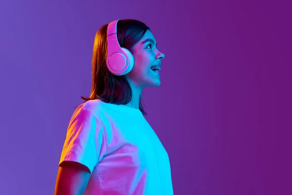 肯定的な若い女の子のサイドビューの肖像画ネオンの光のグラデーション紫色の背景に対してヘッドフォンで音楽を聴く ファッション ライフスタイルの概念 — ストック写真