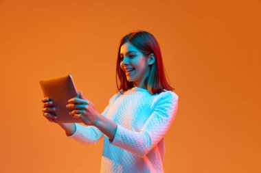 Genç bir kızın portresi, dizüstü bilgisayarda çalışan bir öğrenci, parlak ışıkta turuncu stüdyo arka planına karşı cihazlar kullanıyor. İnsan duyguları kavramı, gençlik, duygular, moda, yaşam tarzı, reklam
