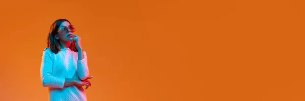 ネオンの光の中でオレンジのスタジオの背景に対して思慮深い顔で立ってカジュアルな服やサングラスの若い美しい女の子 ファッション ライフスタイルの概念 バナー広告 — ストック写真