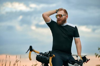Yakışıklı, genç, sakallı, spor gözlüklü, spor kıyafetli, bisikletli arka planda oturuyor. Spor anlayışı, hobi, eğlence etkinliği, eğitim, sağlık, hız, dayanıklılık, reklam