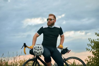 Yakışıklı, genç, sakallı, spor gözlüklü, spor kıyafetli, bisikletli arka planda oturuyor. Spor anlayışı, hobi, eğlence etkinliği, eğitim, sağlık, hız, dayanıklılık, reklam