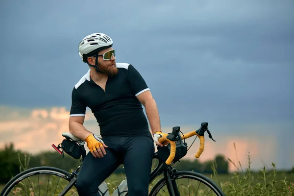 stock image Stylish, bearded, young man in sportswear and helmet, sitting on bike by the road on field in chill summer evening. Concept of sport, hobby, leisure activity, training, health, speed, endurance, ad