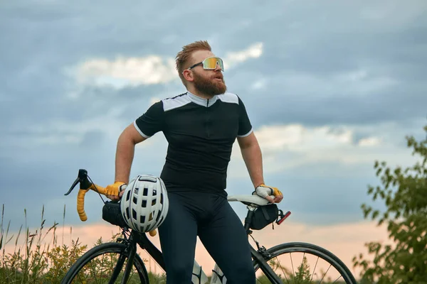 スポーツグラスとスポーツウェアを着たハンサムな若い髭の男が 自転車で畑の背景の上に座っている スポーツ レジャー活動 トレーニング スピード 持久力 広告の概念 — ストック写真