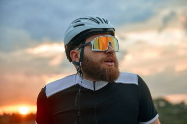 Genç sakallı adamın yakın plan portresi, kasklı bisikletçi ve günbatımı gökyüzü arka planında duran özel gözlükler. Spor anlayışı, hobi, eğlence etkinliği, eğitim, sağlık, hız, dayanıklılık, reklam