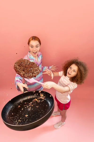 创意形象 在粉红工作室的背景下 小女孩和她年幼的妹妹一起做饭 烤肉当锅子里的汉堡包 生活方式 广告的概念 — 图库照片