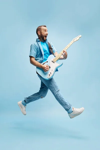 面带微笑的快乐男人 穿着休闲装 在蓝色的工作室背景下愉快地弹奏吉他 业余爱好 广告概念 — 图库照片