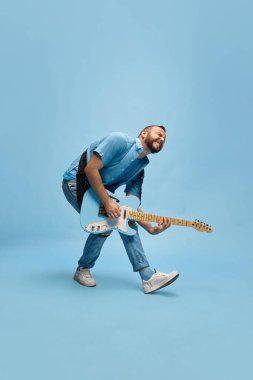Sanatçı, etkileyici, yetenekli, günlük kıyafetler içinde müzisyen mavi stüdyo arka planında elektro gitar çalıyor. Müzik kavramı, yetenek, hobi, eğlence, festival, performans, reklam