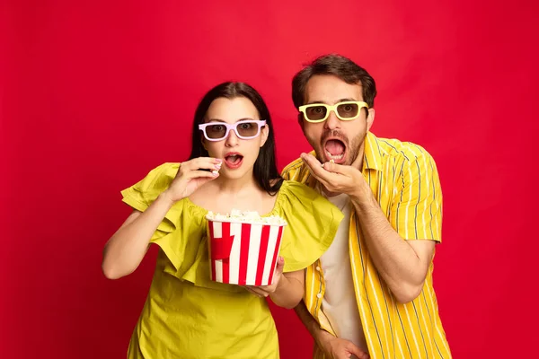 Man and woman emotionally watching movie, eating popcorn against red studio background. Shocked faces. Concept of friendship, relationship, communication, emotions, lifestyle, ad