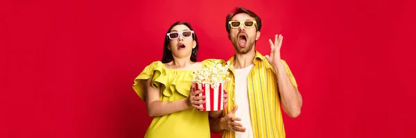 Man and woman emotionally watching movie, eating popcorn against red studio background. Horror movie. Concept of friendship, relationship, communication, emotions, lifestyle, ad