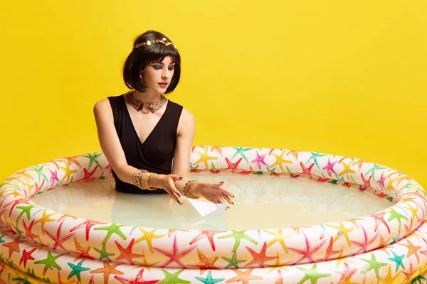 stock image Pretty young woman in image of Cleopatra sitting in swimming pool with milk and playing with paper ship against yellow background. Dreams. Concept of antique culture, history, comparison of eras, art