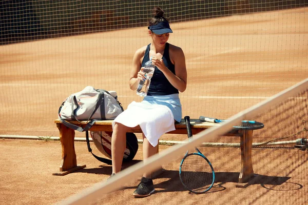 トレーニング ゲーム休息と飲料水の後 屋外テニスコートのベンチに座ってスポーツウェアの若いスポーツウェアの女の子 スポーツ アクティブなライフスタイル 持久力と強さの概念 — ストック写真
