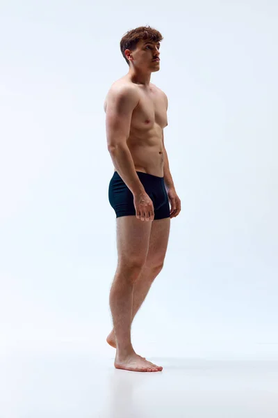 Full-length image of young man with muscular body posing shirtless in underwear against grey studio background. Strength. Concept of mans beauty, sportive and healthy lifestyle, athletic body