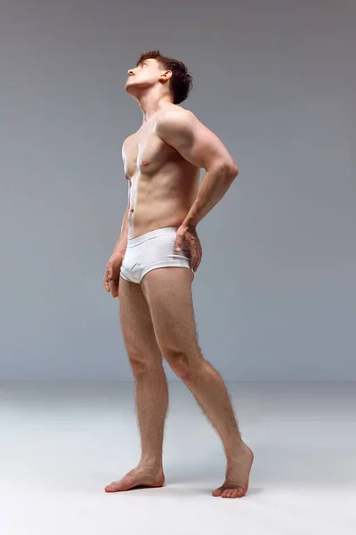 Full-length image of young man with healthy, strong, muscular body posing shirtless in underwear on grey studio background. Back pains. Concept of mans beauty, sport, health, athletic body, medicine