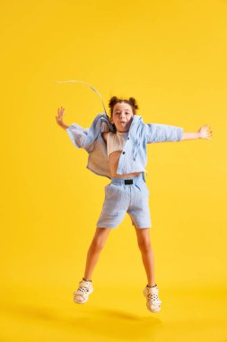 Aktif küçük bir kızın tam boy portresi, günlük giysiler içindeki çocuk sarı stüdyo arka planına neşeyle zıplıyor. Duyguların kavramı, çocukluk, eğitim, moda, yaşam tarzı, reklam