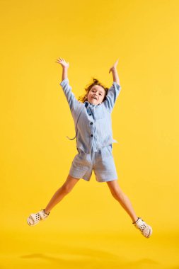 Pozitif, aktif küçük bir kızın tam boy portresi, günlük giysiler içinde sarı stüdyo arka planına neşeyle atlayan bir çocuk. Duyguların kavramı, çocukluk, eğitim, moda, yaşam tarzı, reklam