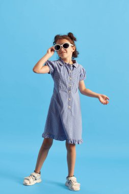 Mavi stüdyo arka planına poz veren şirin elbiseli ve güneş gözlüklü küçük bir kızın tam boy portresi. Duyguların kavramı, çocukluk, eğitim, moda, yaşam tarzı, reklam