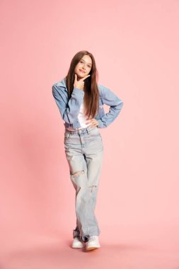 Pembe stüdyo arka planında duran, günlük kıyafetlerle gülümseyen genç, güzel bir kızın tam boy portresi. İnsan duyguları kavramı, moda, gençlik, yaşam tarzı, kadın güzelliği, reklam