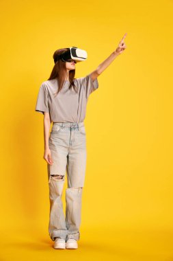Sarı stüdyo arka planında VR gözlük takan, gündelik elbiseli güzel bir kızın tam boy portresi. İnsan duyguları kavramı, moda, gençlik, yaşam tarzı, kadın güzelliği, reklam