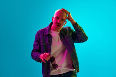 Bıyıklı genç kel adamın portresi neon ışıklarıyla mavi stüdyo arka planına karşı günlük kıyafetler giymiş. Konsantre yüz. İnsan duyguları, yüz ifadesi, yaşam tarzı kavramı