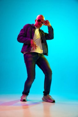 Bıyıklı, sıradan kıyafetler giyen, mavi stüdyo ışıklarında arka planda poz veren kel bir gencin tam boy portresi. İnsan duyguları, yüz ifadesi, yaşam tarzı kavramı