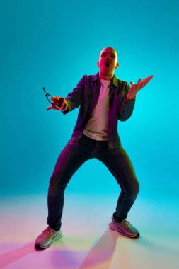 Bıyıklı, sıradan kıyafetler giyen, mavi stüdyo ışıklarında arka planda poz veren kel bir gencin tam boy portresi. İnsan duyguları, yüz ifadesi, yaşam tarzı kavramı