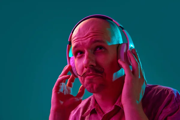 ネオンライトのシアン スタジオの背景を背景にヘッドフォンで音楽を聴く口ひげの若いはげ男の肖像 いい歌だ 人間の感情 ライフスタイルの概念 — ストック写真