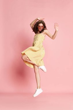 Pembe stüdyo arka planında neşeli bir şekilde dans eden güzel sarı elbiseli ve güneş gözlüklü genç bir kadının tam boy portresi. İnsan duyguları kavramı, moda, güzellik, yaşam tarzı, yaz havası, reklam