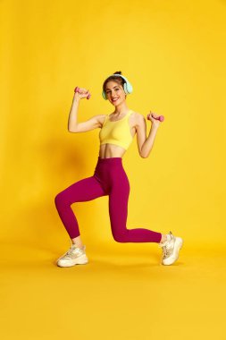 Spor giyim ve çitlerde, sarı stüdyo arka planında dambıllarıyla antrenman yapan zayıf genç bir kadının tam boy görüntüsü. Spor, spor, vücut bakımı, moda, gençlik, yaşam tarzı, reklam