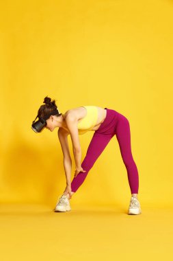 Gözlük takan ve sarı arka planda eğitim gören zayıf bir genç kadının tam boy görüntüsü. Isınma, ısınma. Spor, spor, vücut bakımı, moda, gençlik, yaşam tarzı, reklam