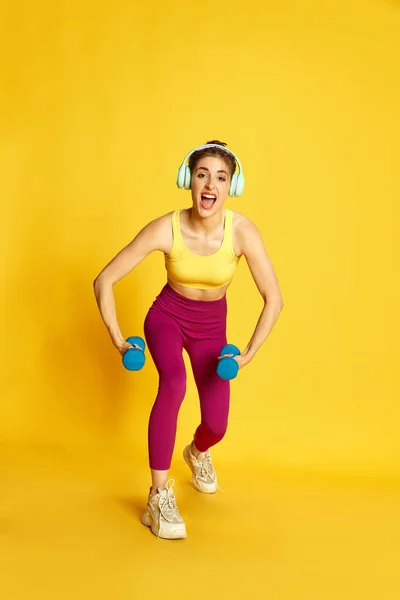 若いスポーツのフル長さの画像 ヘッドフォンのトレーニングでスリムな女性 黄色の背景に対するダンベルで演習を行います スポーツ フィットネス ボディケア ファッション ライフスタイル 広告の概念 — ストック写真