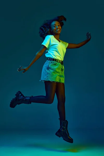 穿着休闲装的快乐 年轻的非洲女人的画像 在霓虹灯下 带着微笑在青色 蓝色的工作室背景下跳跃 生活方式 广告的概念 — 图库照片