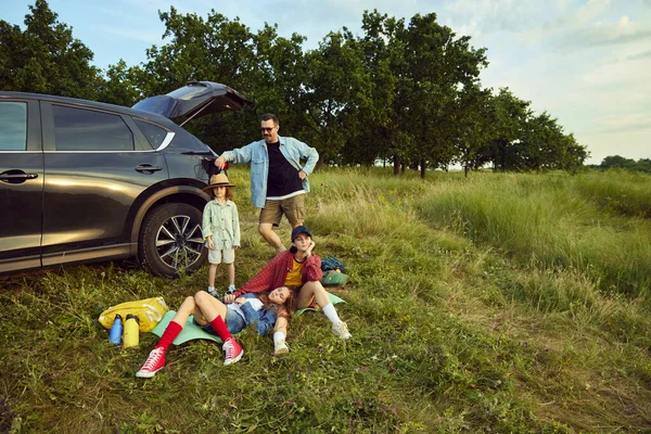 stock image Man, woman and children sitting by the car on fitness matt and resting after active hiking on hills. Family gathering. Concept of leisure time, fun, nature, active lifestyle, family, travel