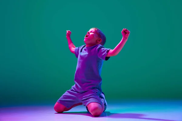 动情的孩子 小男孩高高地坐在膝盖上 在霓虹灯下 在青色的工作室背景下表现出胜利的姿态 生活方式 广告的概念 — 图库照片