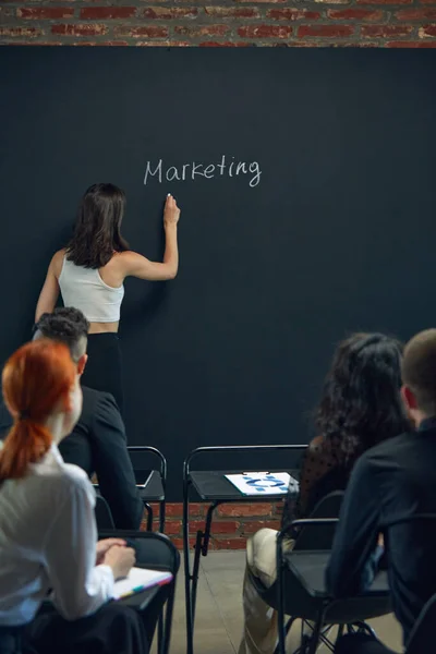 Young woman, employee standing by blackboard and writing word of marketing. Meeting with colleagues. Discussing on projects. Concept of business, planning, strategy, brainstorming, analytics, teamwork