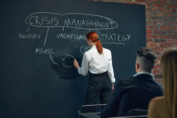 Young woman, employee writing on black board plan of crisis management. Meeting with colleagues. Development of successful working system. Concept of business, strategy, brainstorming, analytics, ad