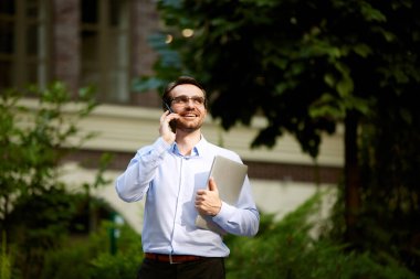 İş adamı ofisin dışında yürüyor, dizüstü bilgisayarını tutuyor, cep telefonuyla konuşuyor. Anında çalışıyor. İş kavramı, kariyer gelişimi, hırslar, başarı, ofis yaşam tarzı