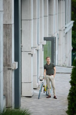 Yakışıklı, şık, şık giyimli genç iş adamı modern binaların yanında bisikletiyle dikiliyor. İşe gidiyorum. İş kavramı, aktif yaşam tarzı, moda, gençlik, ekoloji