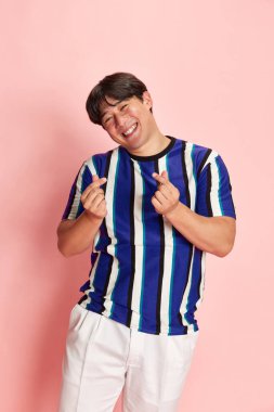 Gülümsemesi ve pozitifliği olan sıradan giyinmiş genç Koreli adam pembe stüdyo geçmişine karşı bir jest yapıyor. İnsan duyguları kavramı, yaşam tarzı, gündelik moda, pozitiflik, reklam