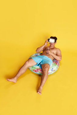 Yüzme şortlu ve video gözlüklü genç bir adam, sarı stüdyo arka planında yüzme çemberinde oturuyor. Yaz kavramı, tatil, boş zaman, tatiller, insan duyguları, reklam