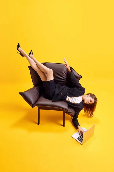 不快な立場でアームチェアに横たわる若いビジネス女性は 明るい黄色の背景に対してラップトップに取り組んでいます ビジネス ワーキングルーチン 締め切り フリーランス オフィス — ストック写真