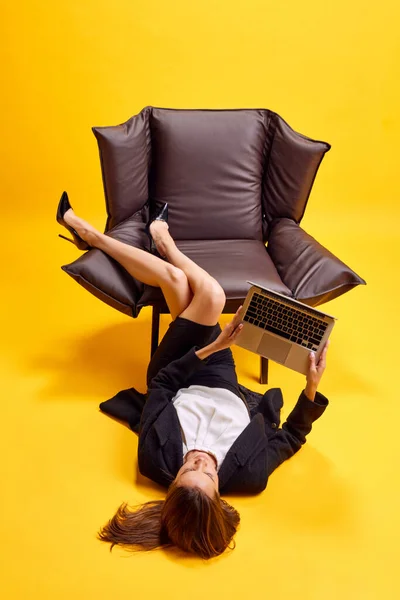 若いビジネス女性 従業員は椅子の近くの床に横たわり 明るい黄色の背景に対してラップトップでオンラインで作業しています ビジネス ワーキングルーチン 締め切り フリーランス オフィス — ストック写真